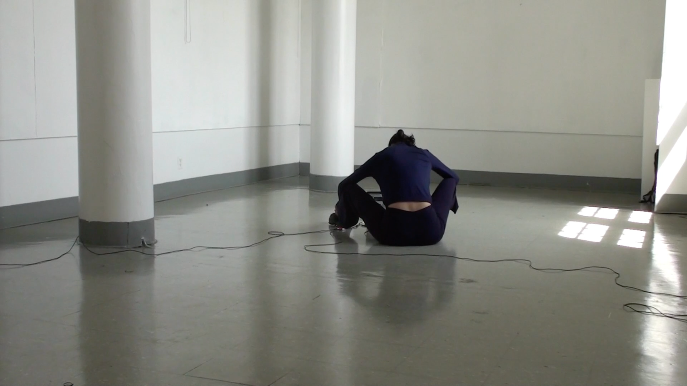 Performance still, performer sits in the center of the room with their back to the camera, cables run out from the performers body towards the camera view and out of the frame.